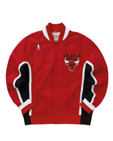 A red Chicago Bulls basketball jacket with black and white striped sleeve panels, featuring the Chicago Bulls logo on the chest and a small NBA logo on the left sleeve.